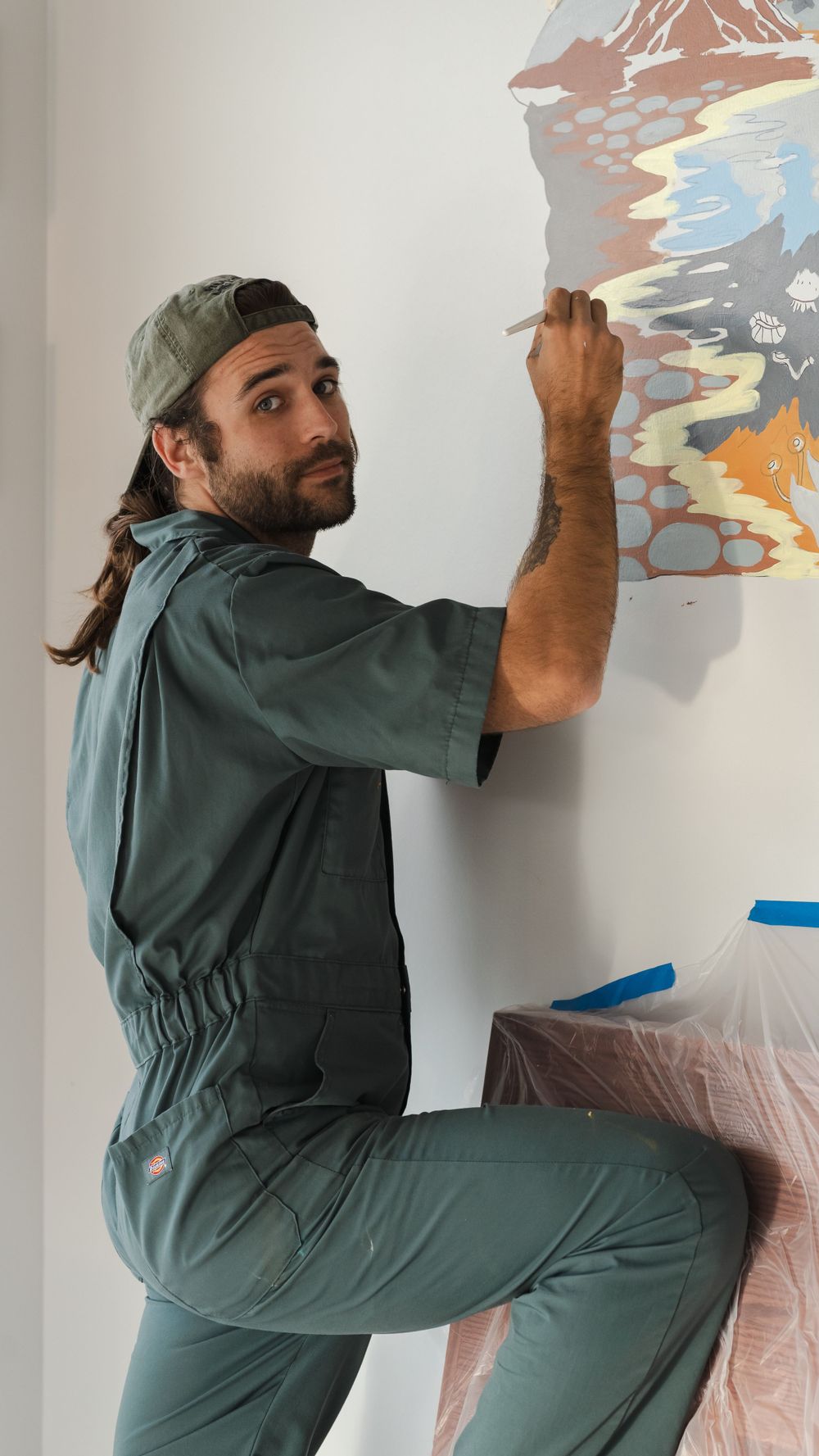 Artist in green overalls working on painting large wall mural.