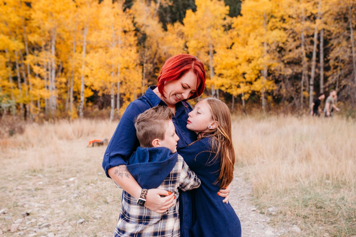 A tender family moment captured in a field with vibrant fall foliage behind them.