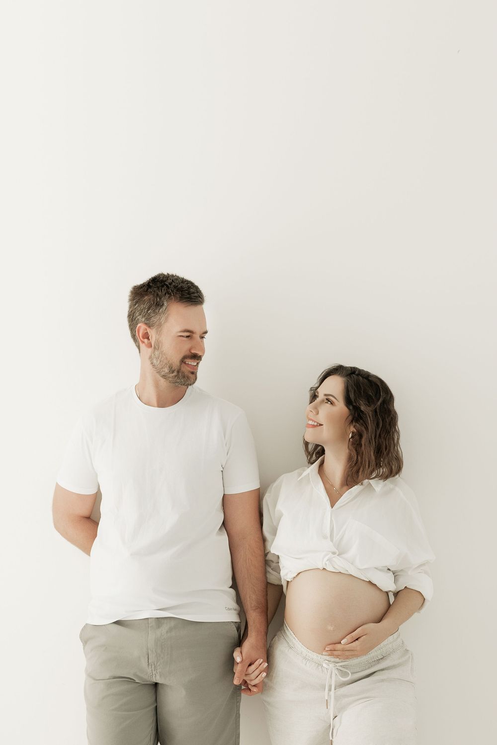 Minimalist maternity photo series showing couple in white and beige against plain wall in three intimate poses.
