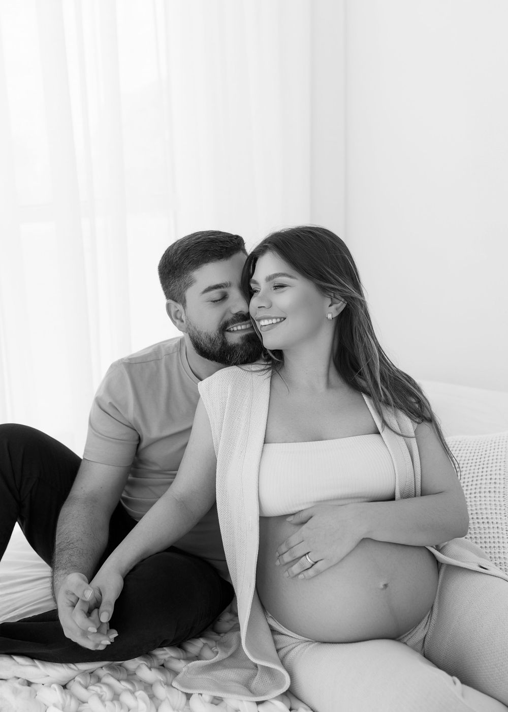 A black and white maternity photo series showing intimate moments between an expecting couple on a white bed.