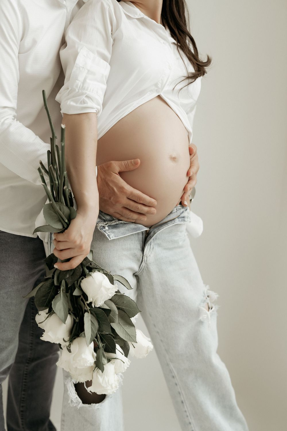 Romantic maternity photo with white roses and casual attire against light background.