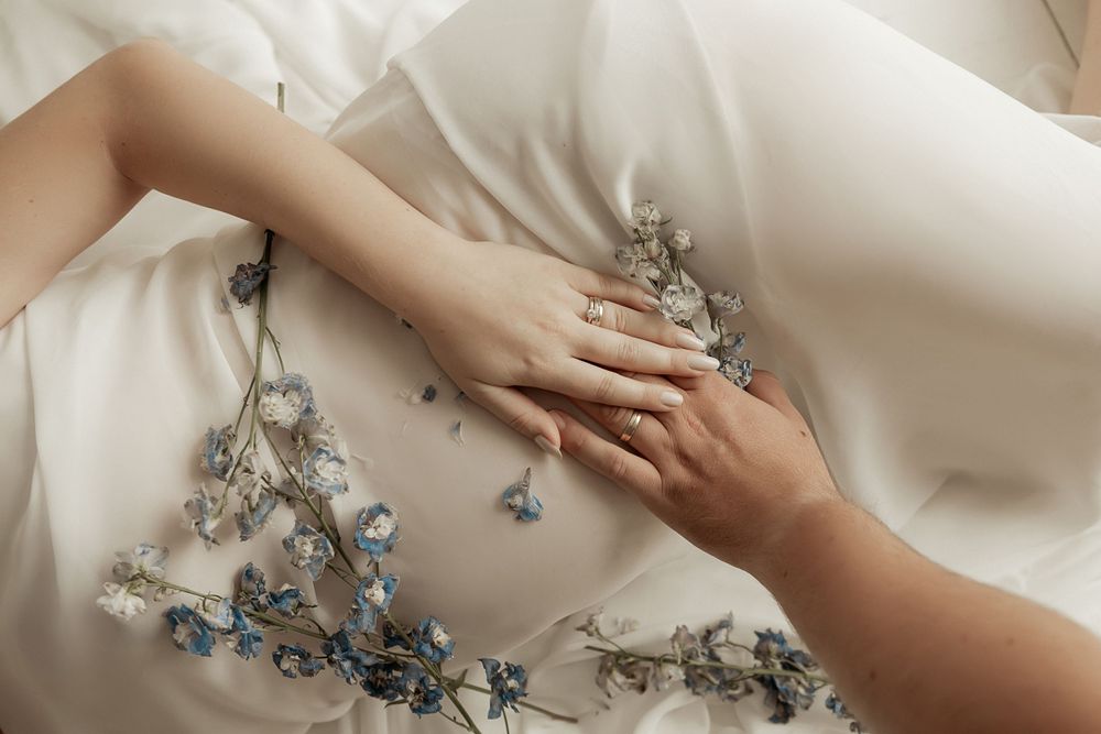 Two delicate hands rest on white fabric adorned with blue floral embroidery and silver threading.