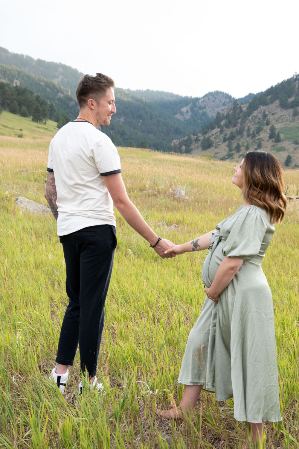 A couple walks hand in hand through tall grass with mountains in the distance.