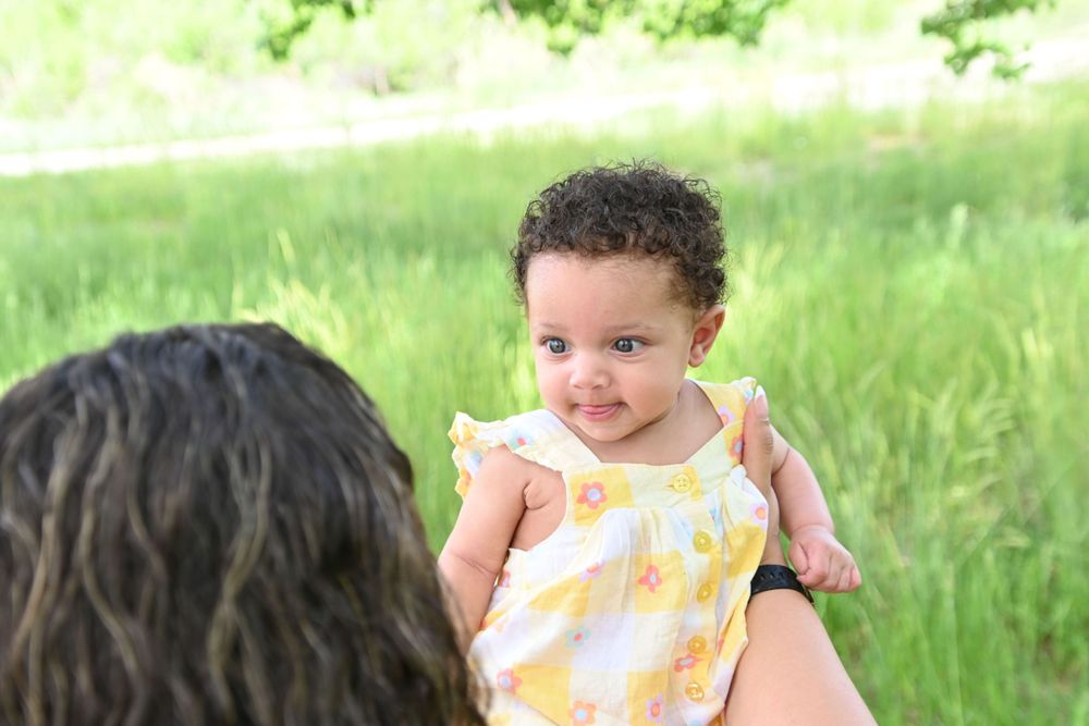 A baby in a yellow dress smiles while being held up outdoors against a blurred natural background.