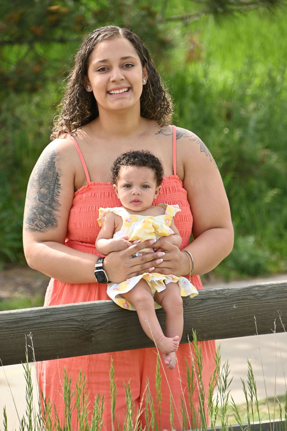 A parent in a coral dress holds a baby in yellow while standing by a wooden fence outdoors.