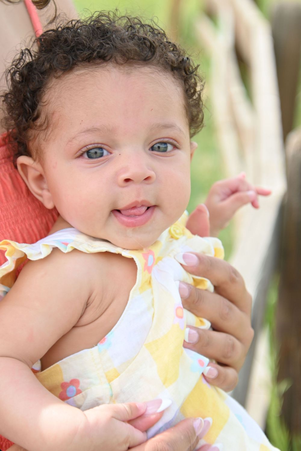 A close-up portrait of a baby with curly hair wearing a yellow and white floral outfit.