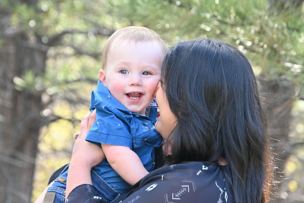 A parent holds a smiling baby wearing blue against an autumn forest backdrop.