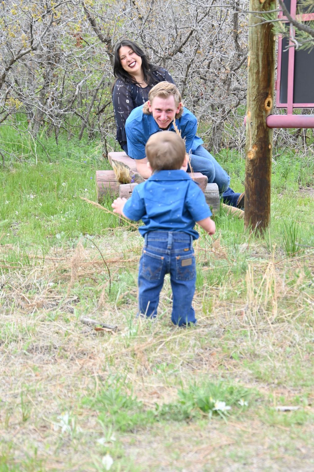 A playful family moment captured outdoors with a toddler in blue denim and adults on playground equipment.