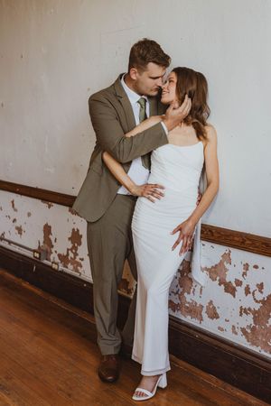 Couple in wedding attire embracing against rustic wall, groom in olive suit kissing bride in white gown.