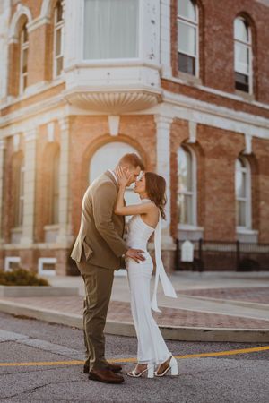Couple in wedding attire sharing a romantic moment at golden hour outside a historic brick building.