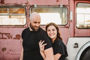 A couple poses together in front of a vintage pink bus showing off an engagement ring.