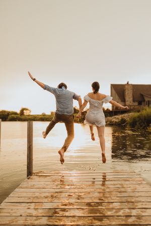 Playful couple jumping off wooden dock at sunset.