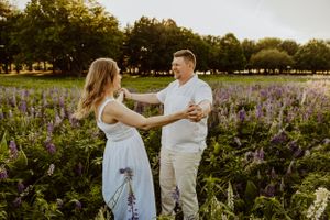 Romantic couple in white outfits posing in a field of purple wildflowers at sunset.
