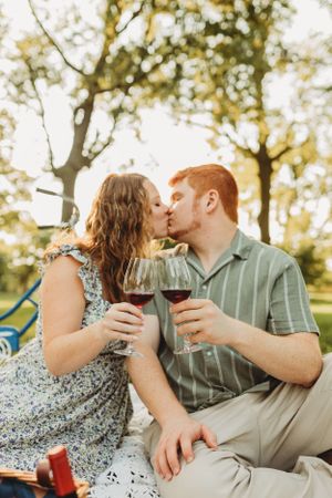 Romantic picnic scene with couple sharing wine glasses and kiss under trees.