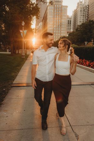 Couple walking together at sunset on city sidewalk with skyscrapers and flowers in background.