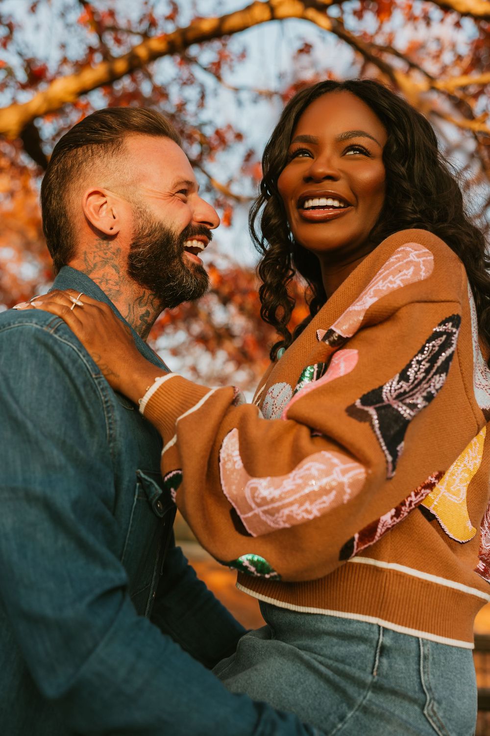 Couple laughing together outdoors with glowing orange fall foliage in background.