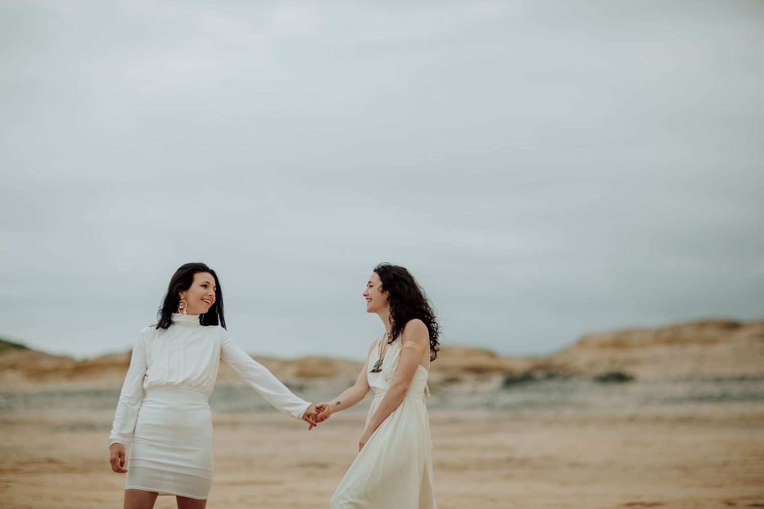 Two people in white dresses hold hands on a rocky beach against an overcast sky Cornish elopement photography beach wedding