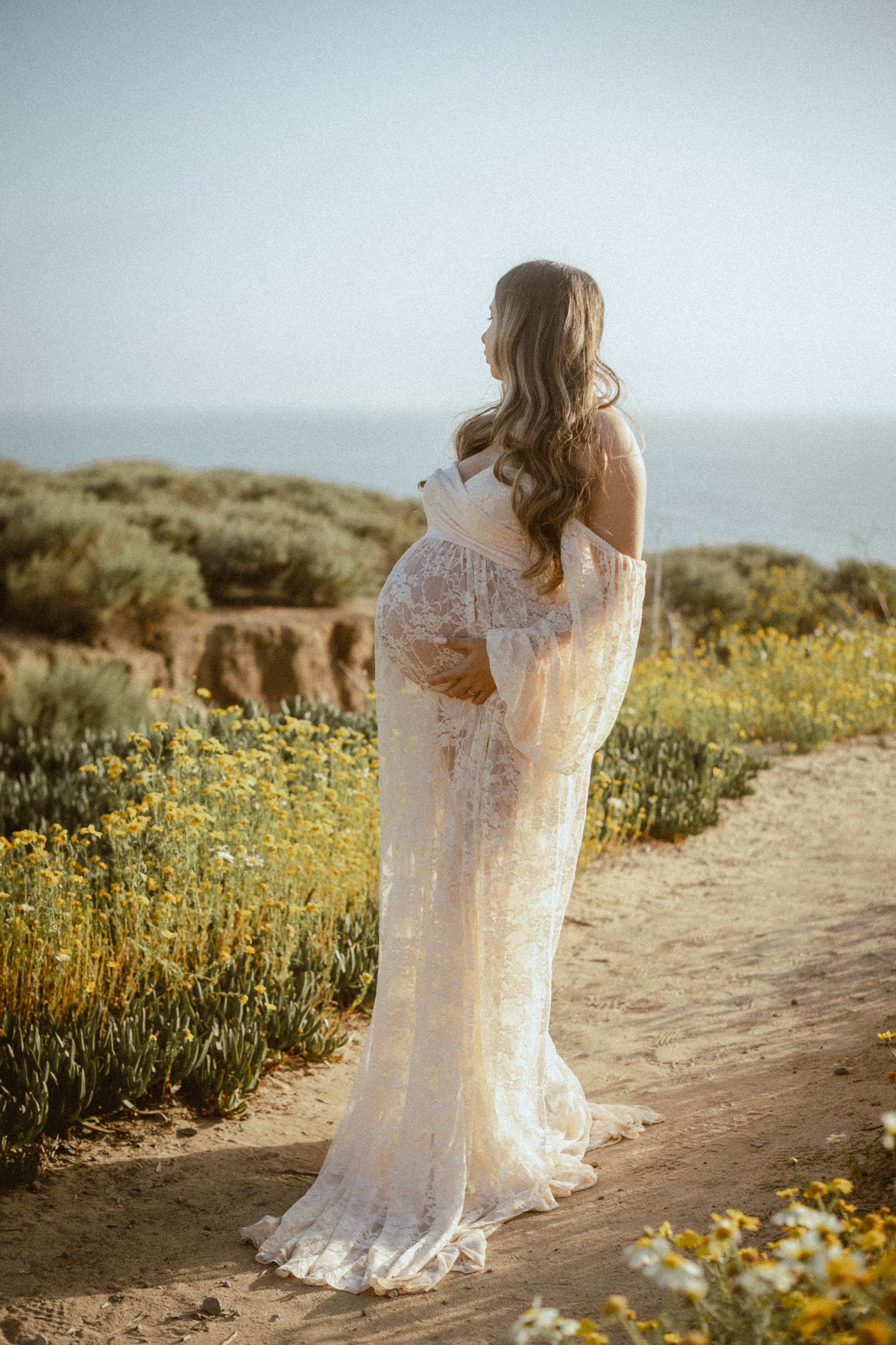 Person in lace dress poses among yellow wildflowers on coastal path at sunset.