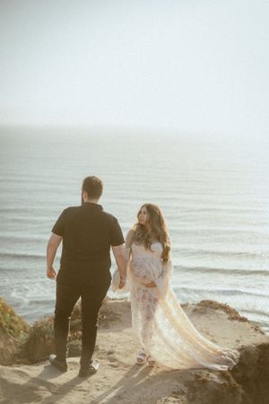 Couple walking along a misty beach at sunset, holding hands with flowing dress trailing behind.