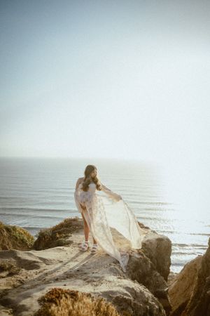 Romantic seaside photo shoot with flowing white lace dress against ocean backdrop.
