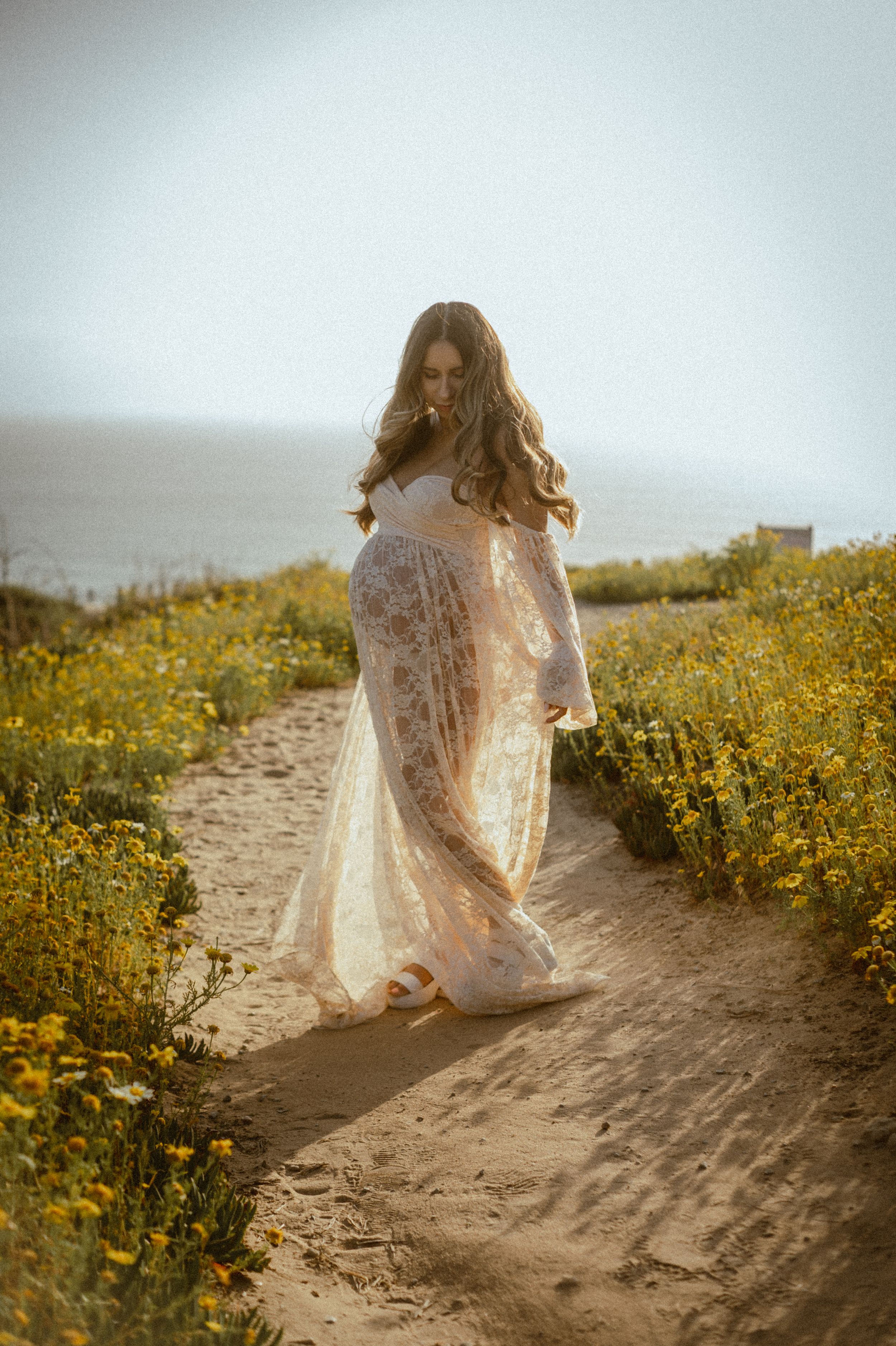 Figure in flowing white lace dress walks through field of yellow wildflowers.