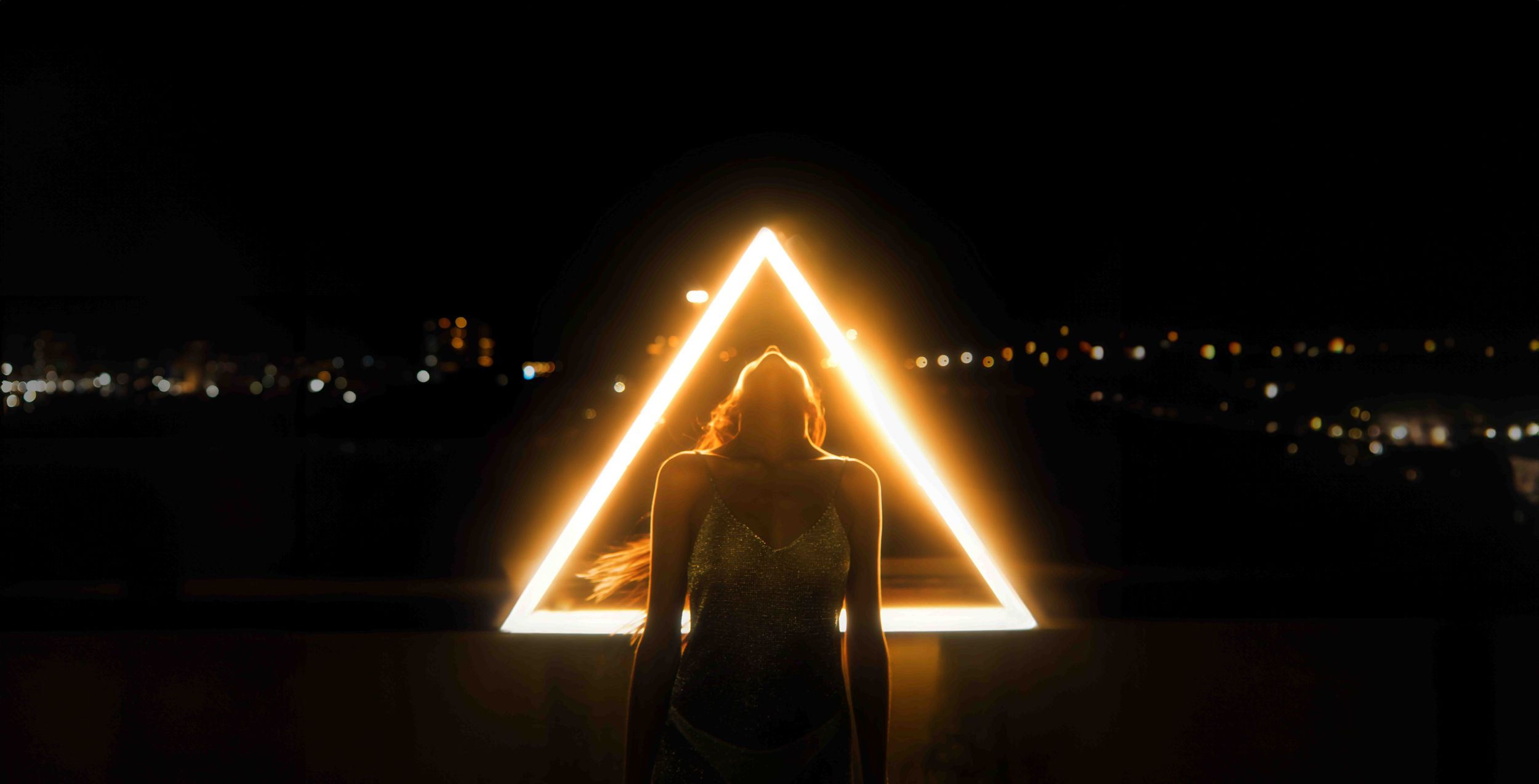 Silhouette standing in front of glowing neon triangle at night.