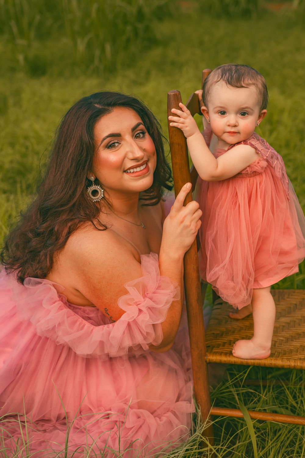 Mother and baby in matching pink tulle dresses pose with wooden chair in outdoor garden setting.