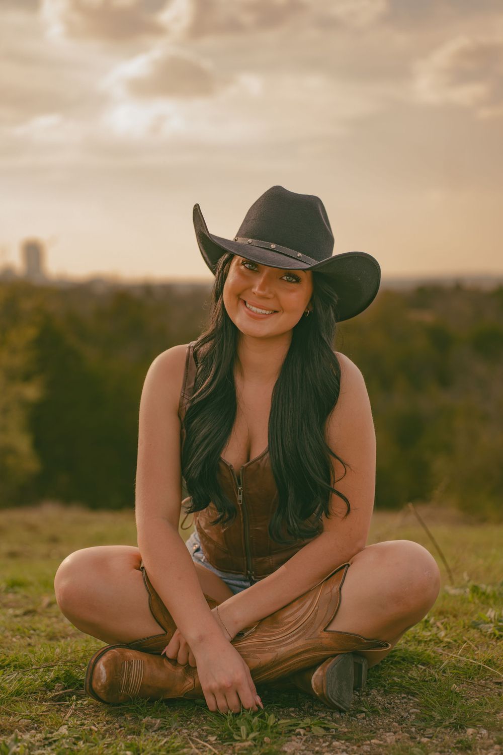 Woman in cowboy hat and boots sits cross-legged on hilltop during golden hour.