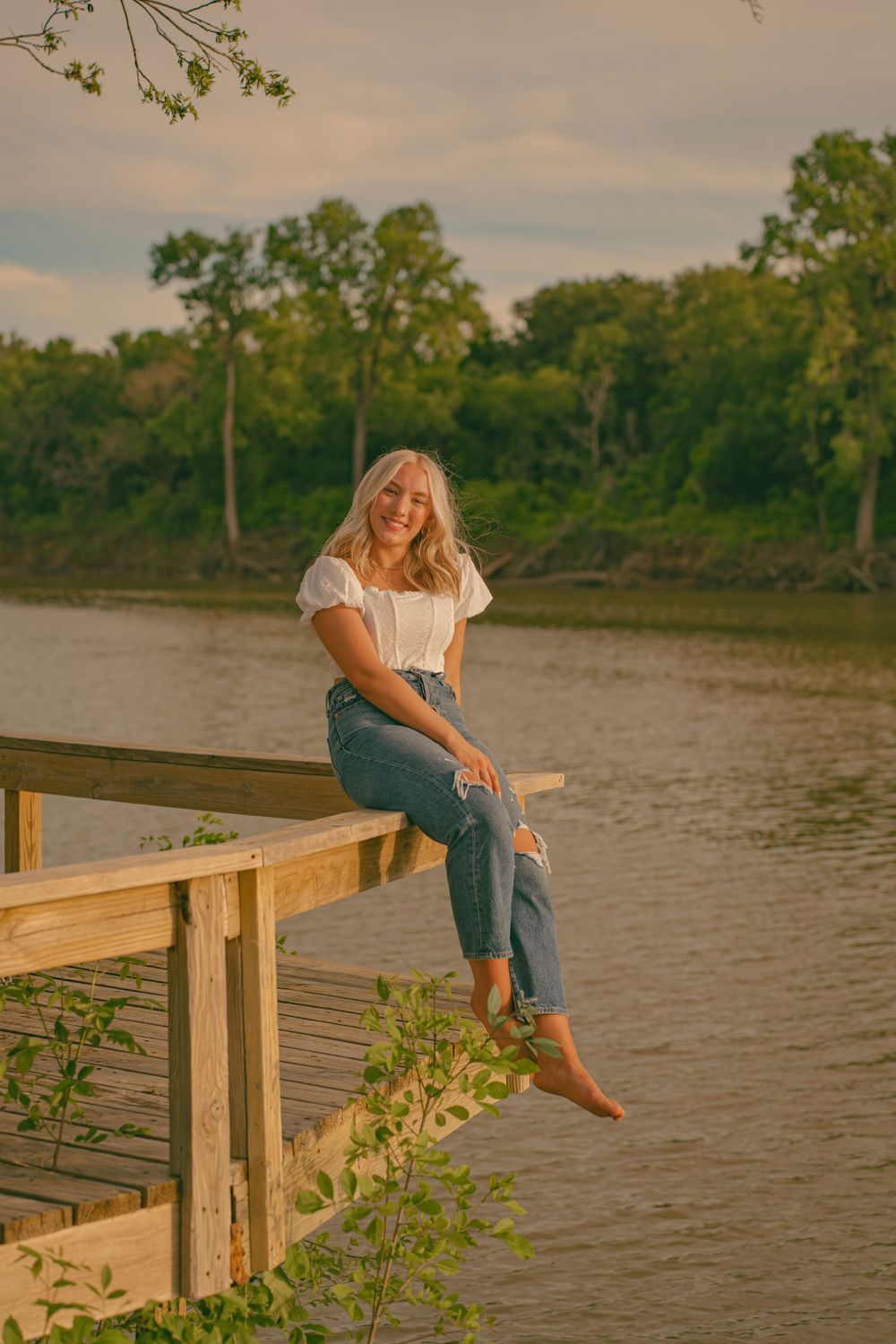 Woman in casual attire sits on wooden dock overlooking river at sunset.