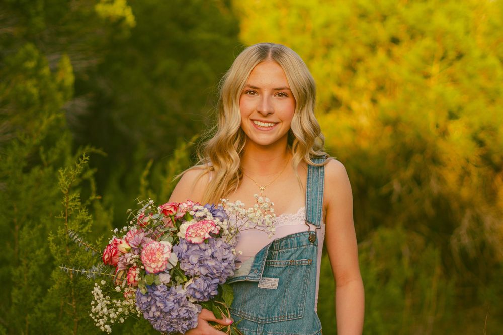 A person in overalls holds a vibrant bouquet of pink and purple flowers against a golden sunset background.