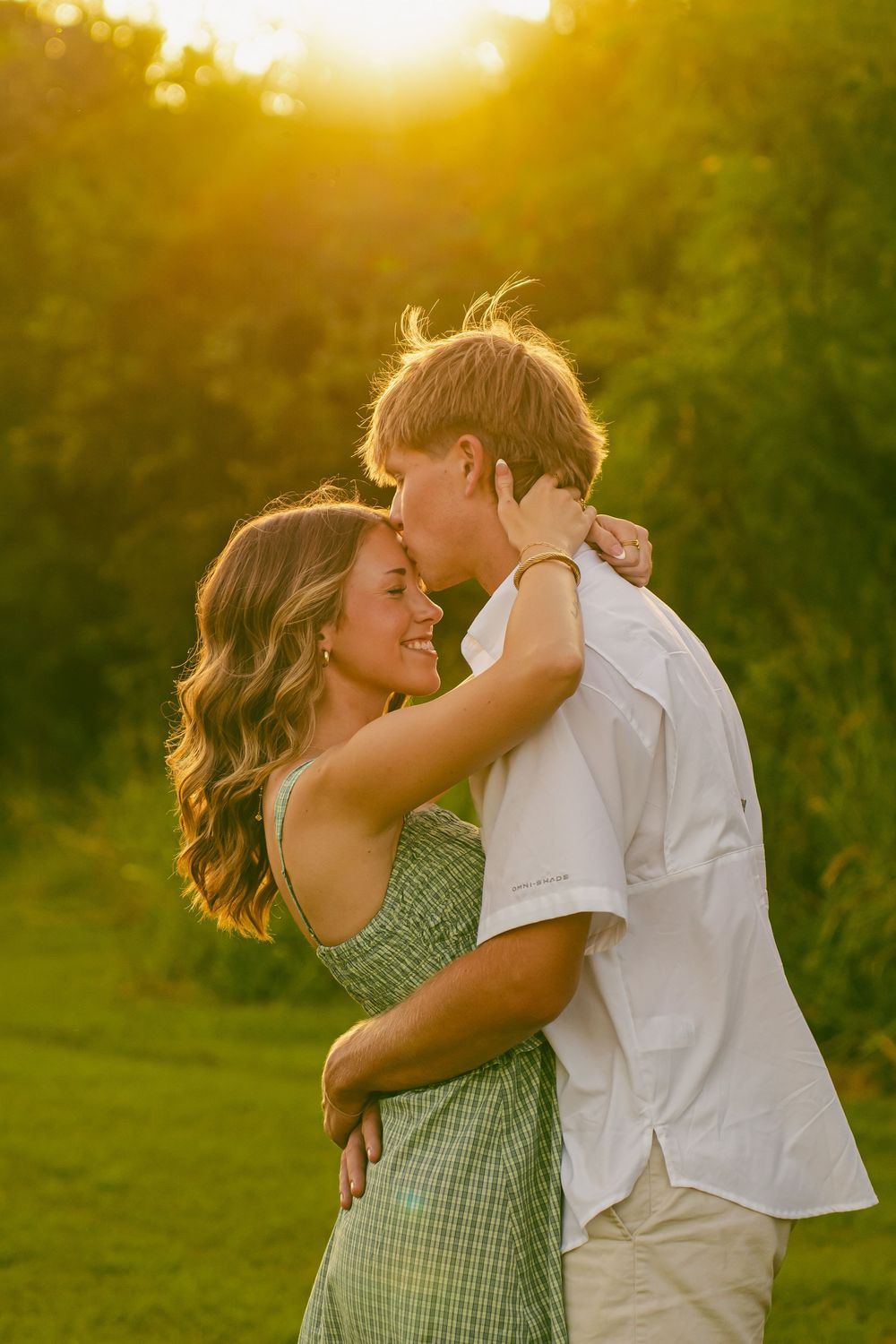 A young couple embraces at golden hour in a lush green field, smiling warmly in a romantic outdoor portrait.