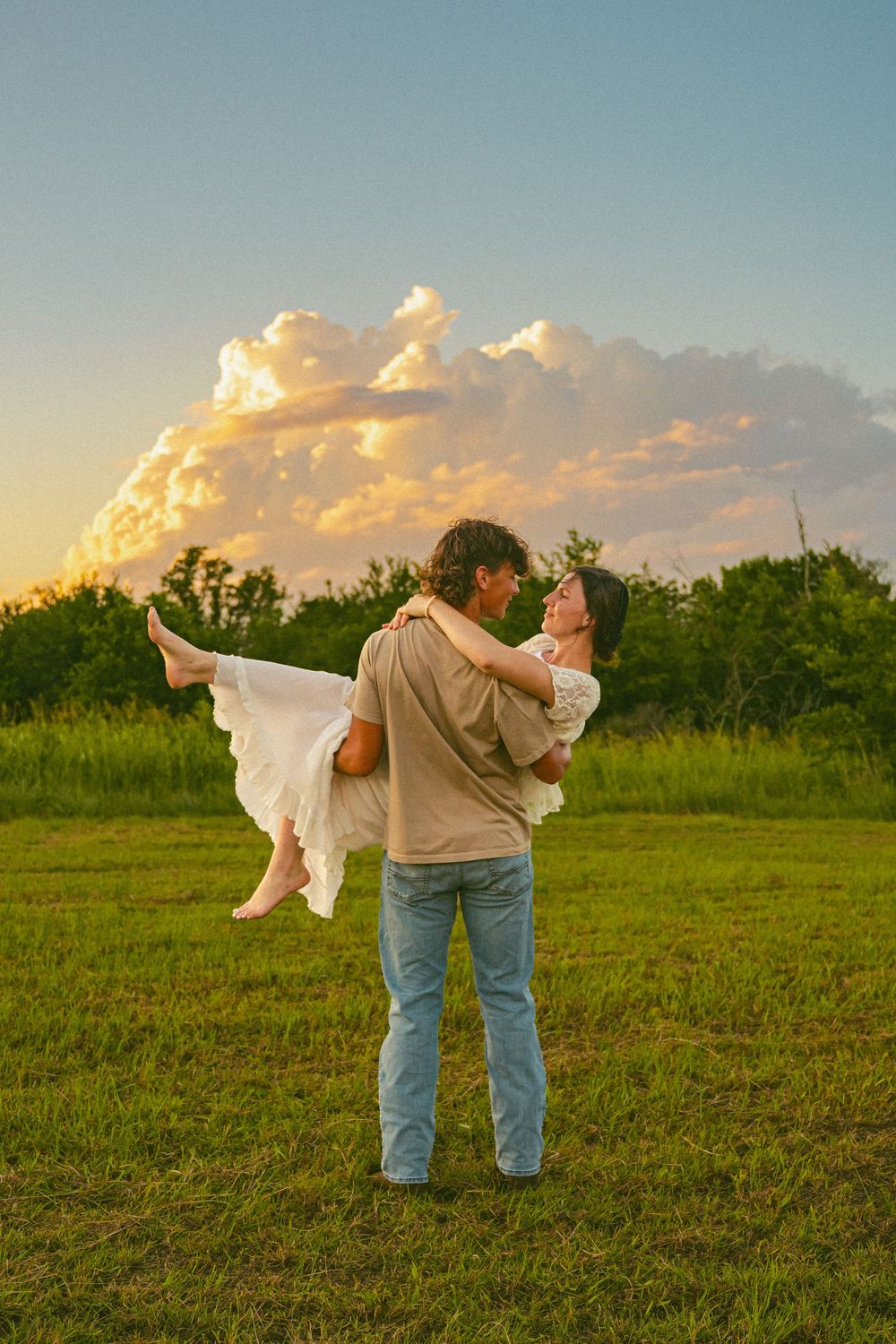 Man carrying woman in white dress in a green field at sunset with dramatic clouds in the background.