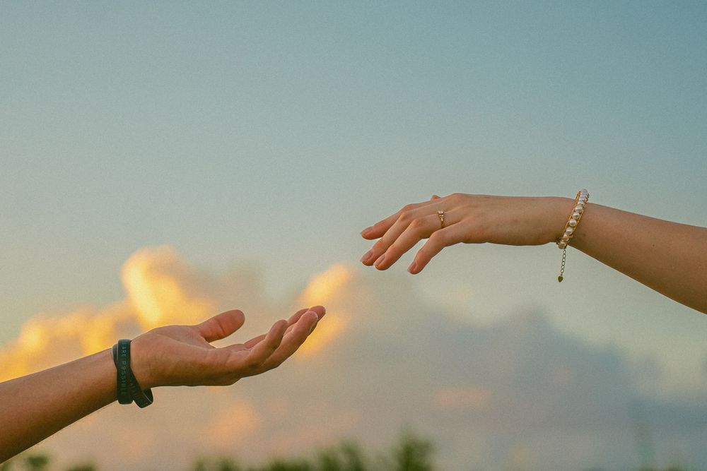 Two hands reaching toward each other at sunset, one wearing a bracelet, against a golden cloudy sky.