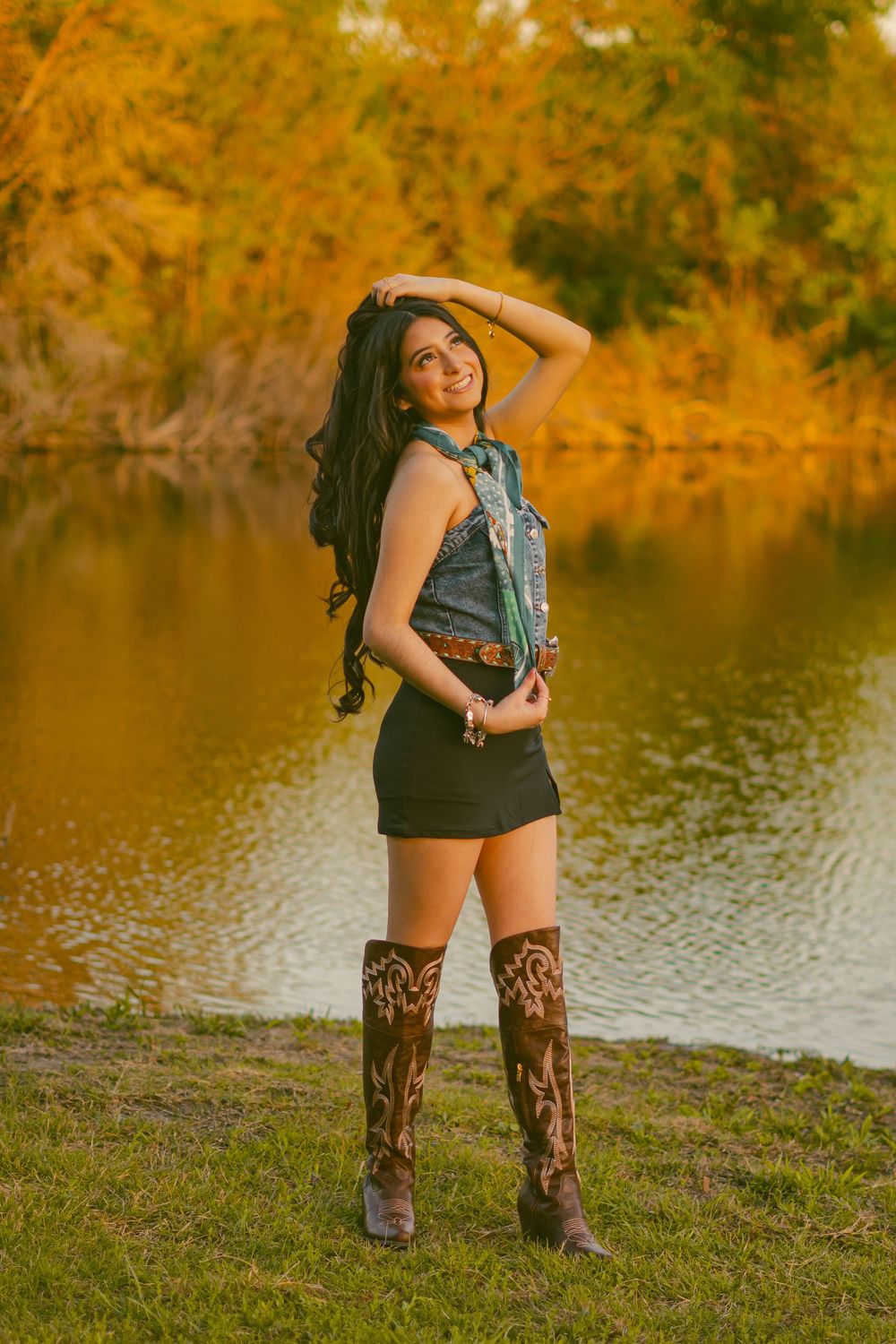 A person in denim vest and tall boots poses by a lake with autumn foliage in the background.