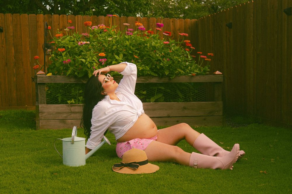Pregnant woman relaxing on grass in a sunny backyard garden with flowers, wearing pink shorts and white shirt.