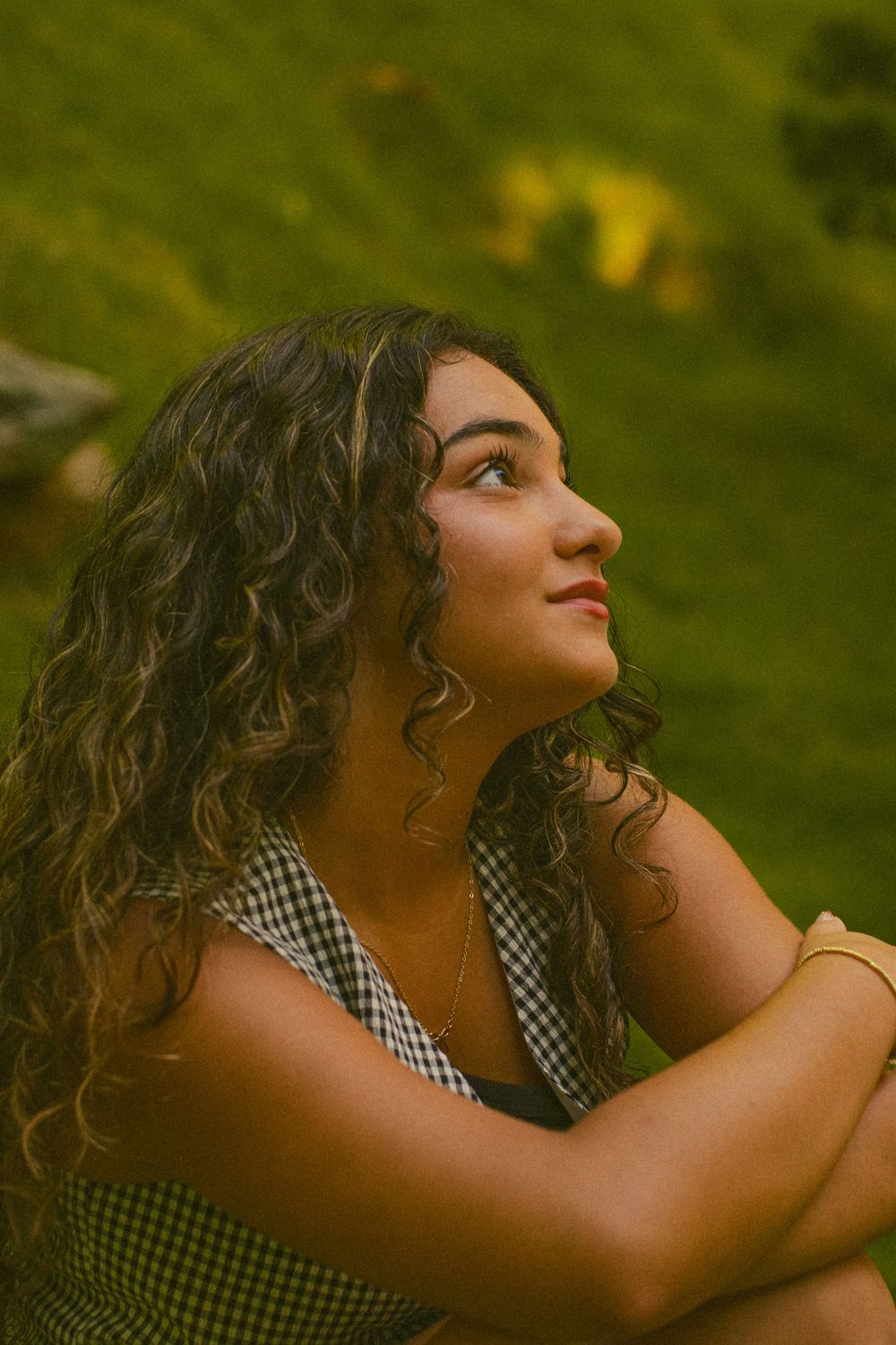 Young woman with curly hair sitting outdoors on green grass, gazing upward thoughtfully in natural light.