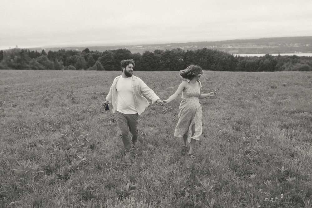A couple holds hands while running through a scenic meadow with rolling hills in the background.