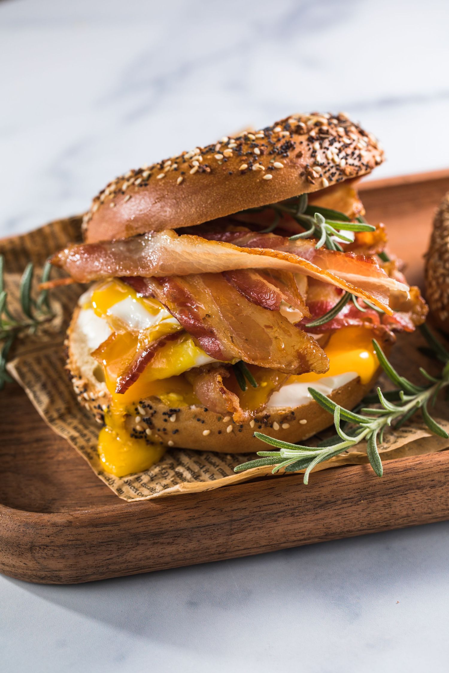 Toasted everything bagel sandwich with crispy bacon, fried egg, and cheese on a wooden board with rosemary.