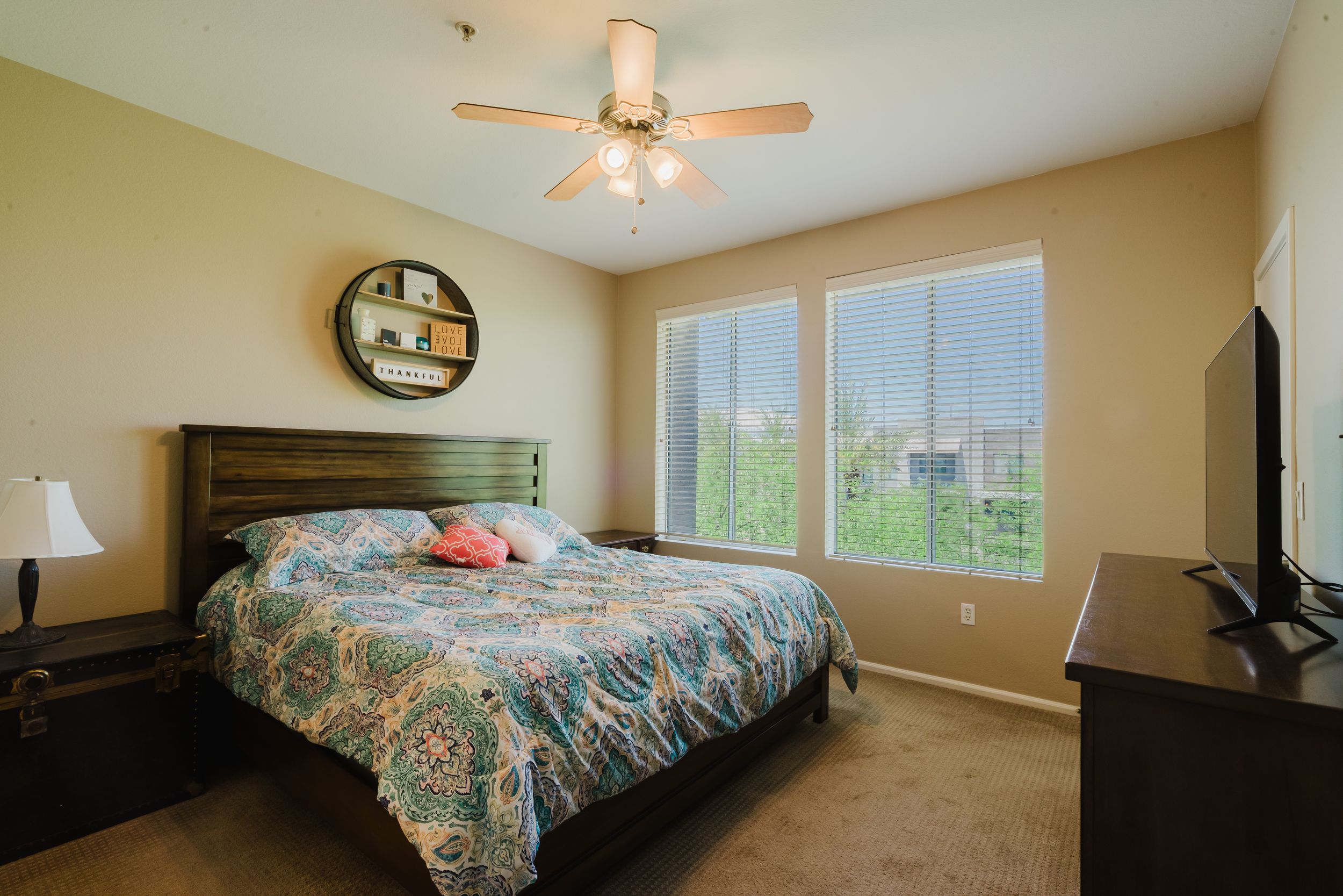 Cozy bedroom with floral bedding, dark wood furniture, ceiling fan, and large windows offering natural light.