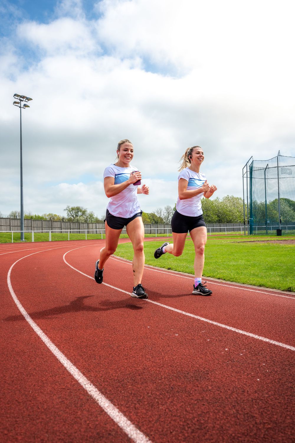 Two athletes perform dynamic stretching exercises on a red running track during outdoor training session.