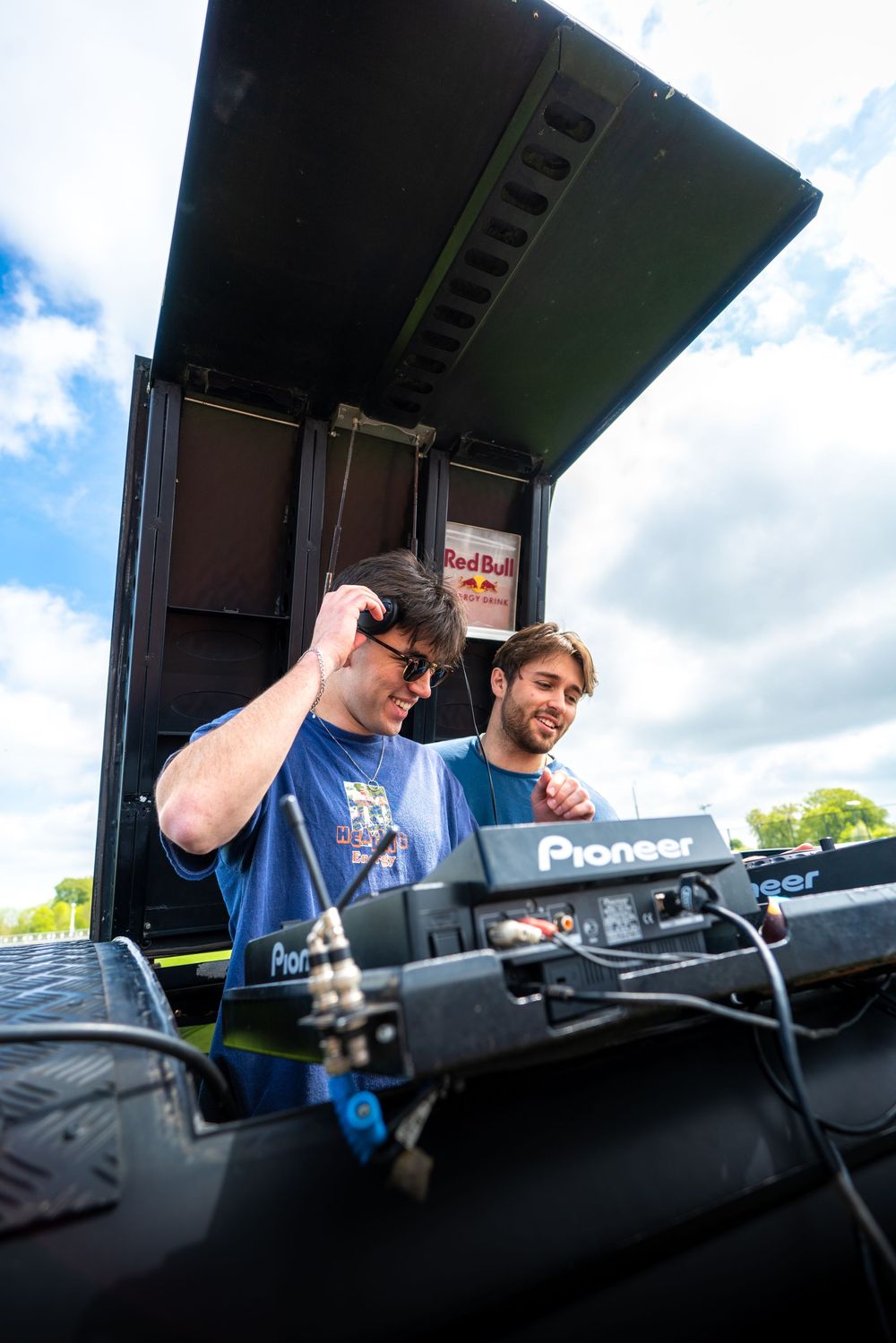 DJs perform at an outdoor music festival behind Pioneer DJ equipment under a cloudy sky.
