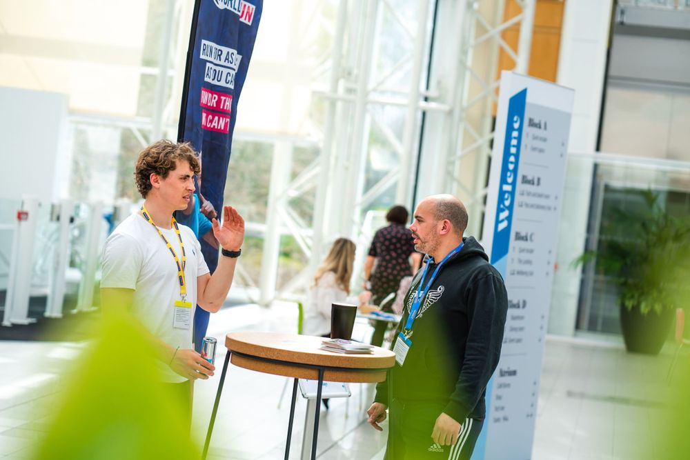 Two people engage in conversation at a professional conference registration table beside informational banners.
