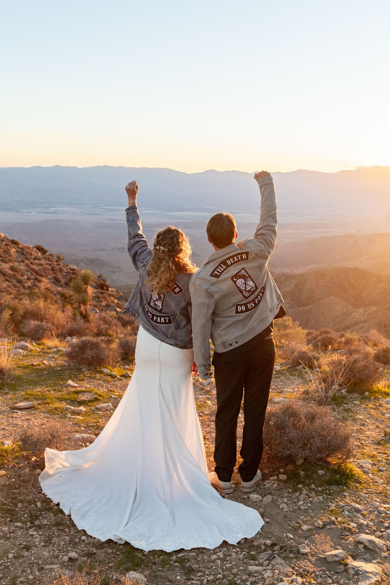 Bride and groom in wedding attire and matching biker jackets raise fists at a stunning mountain sunset overlook.