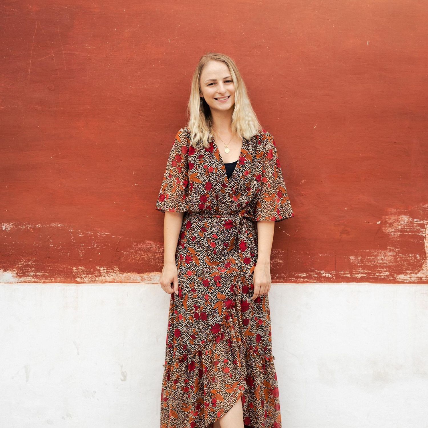 A woman in a floral print dress poses against a red and white wall background.