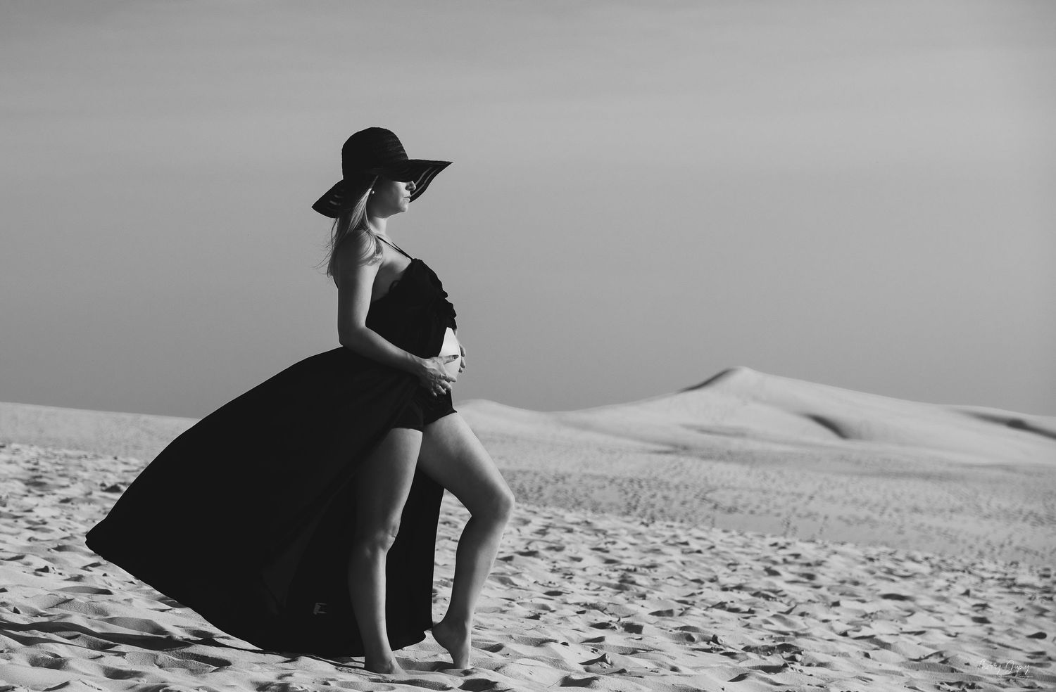 Silhouette d'une femme en robe fluide assise sur les dunes sous un ciel gris