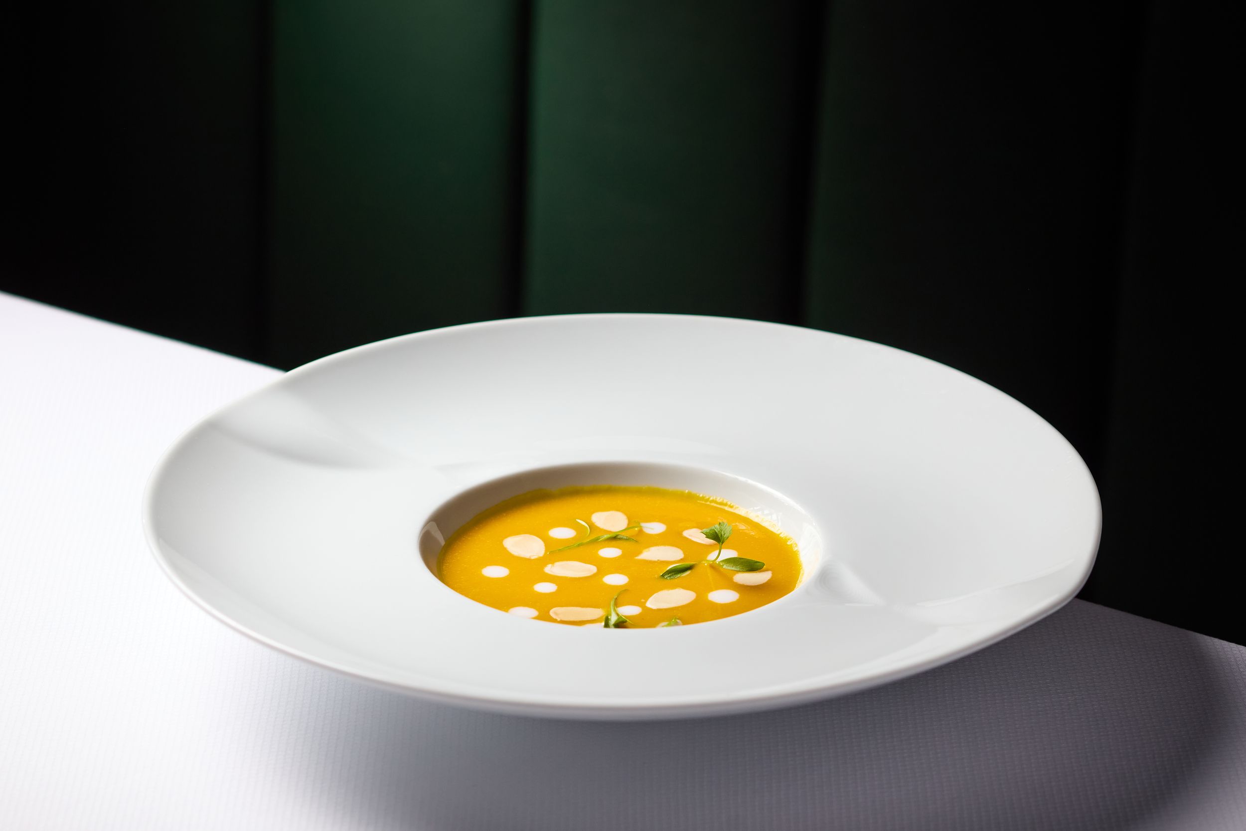Elegant white bowl with creamy orange pumpkin soup garnished with cream dots and herbs on a white table.