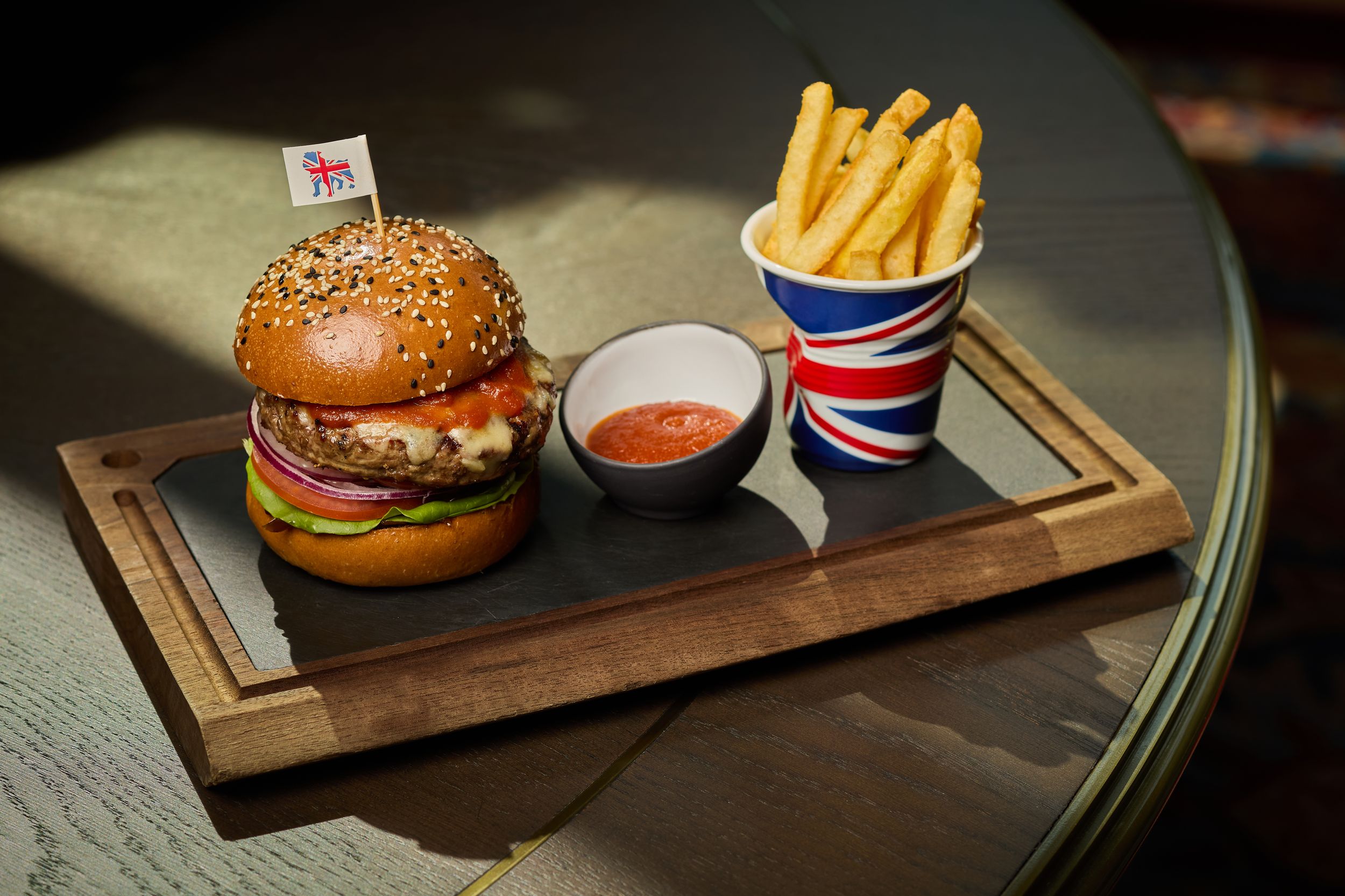 Gourmet burger with sesame bun served with fries and dipping sauce on wooden tray.