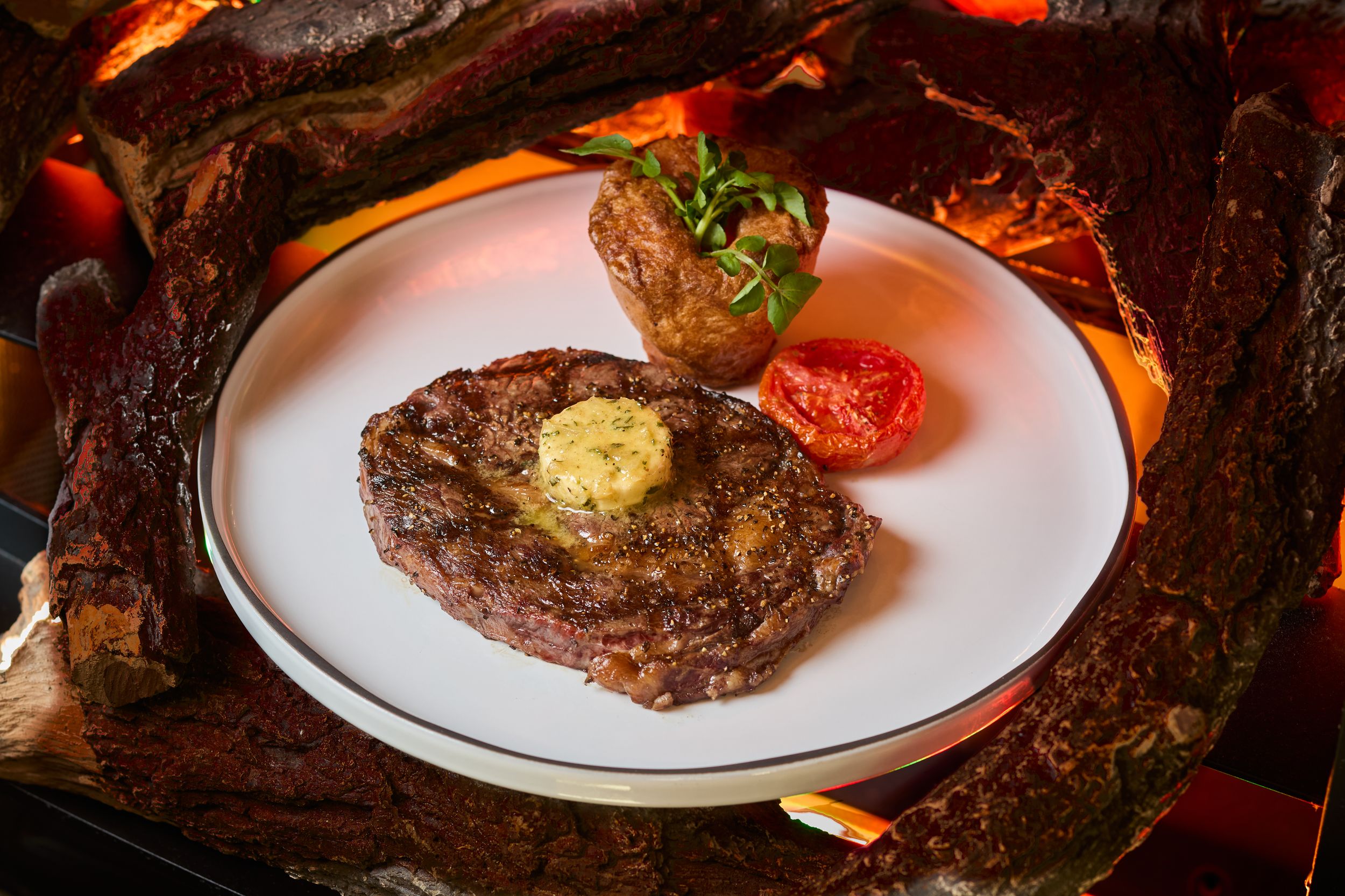 Juicy grilled ribeye steak topped with herb butter, served with a baked potato and tomato on a white plate.