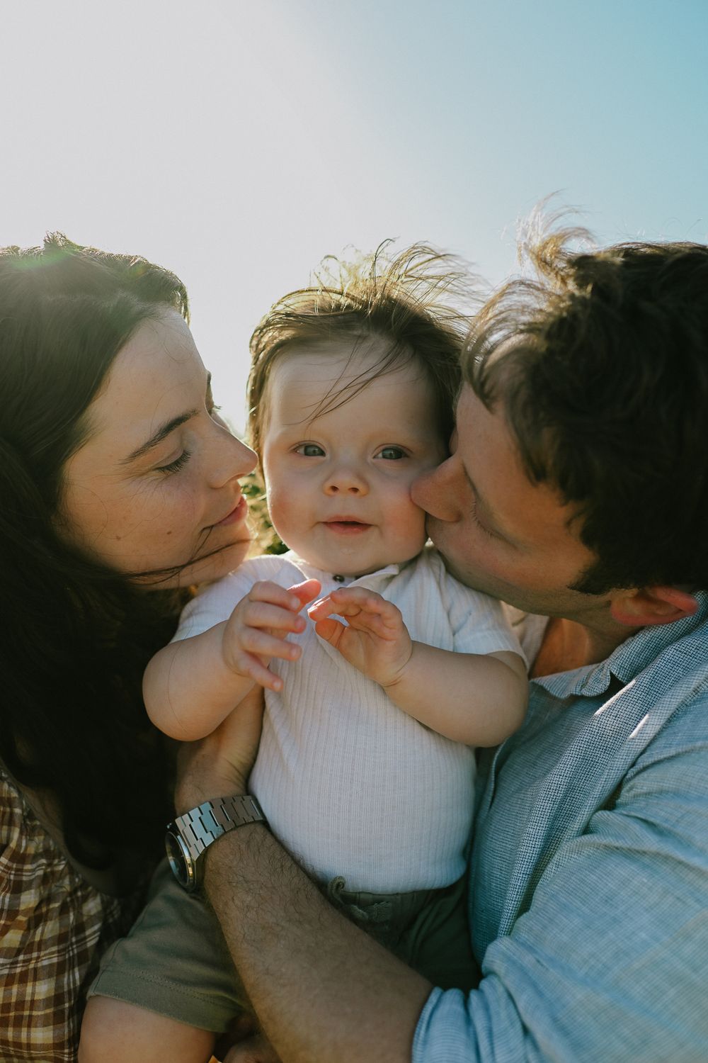 A family shares tender kisses and cuddles together outdoors in warm sunlight.