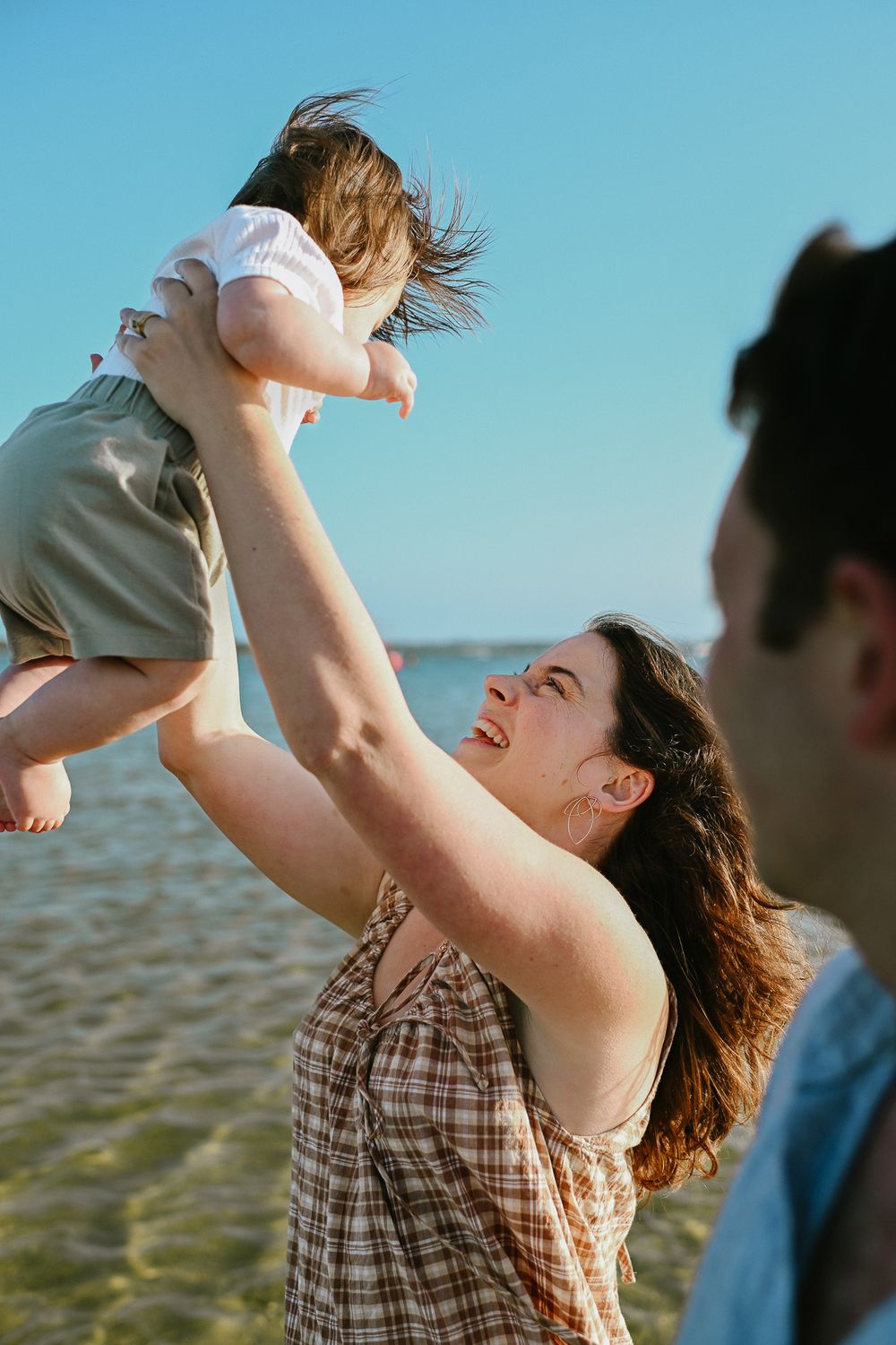 A person lifts a small child up against a bright blue sky near the ocean.